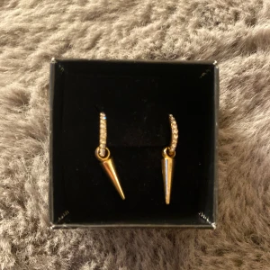 Guldiga örhängen med spetsig hängande detalj - Säljer ett par trendiga örhängen i guld med spetsig hängande detalj. Örhängena har små gnistrande stenar på bygeln som ger extra glans. Perfekta för att lyfta din look!