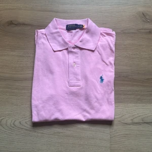 Rosa pikétröja från Polo Ralph Lauren - Säljer en klassisk rosa pikétröja från Polo Ralph Lauren i storlek M med blå broderad logga på bröstet. Tröjan har korta ärmar, krage och knäppning med två knappar. Perfekt för en stilren look. I ny skick och nästan oanvänd. Pris kan diskuteras vid snabb affär.