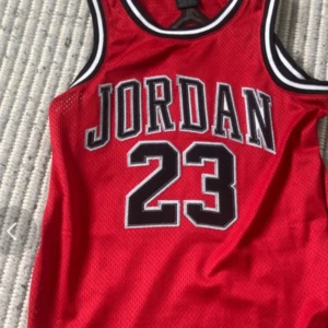 Röd Jordan basketlinne nr 23 - Säljer en röd basketlinne från Jordan med nummer 23 och stora svarta och vita bokstäver framtill. Klassisk sportig look med meshmaterial och svarta/vita detaljer runt hals och ärmhål. Perfekt för dig som gillar streetwear eller basket.