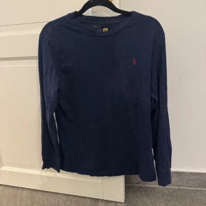 Mörkblå långärmad tröja Polo Ralph Lauren - Snygg mörkblå långärmad tröja från Polo Ralph Lauren med klassisk röd logga broderad på bröstet. Tröjan har rund halsringning och är gjord i mjuk bomull, perfekt för en stilren och avslappnad look.