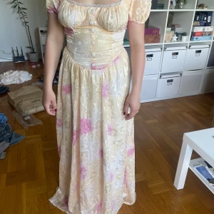 Ljus långklänning med blommigt mönster - Romantisk långklänning i ljusa toner med rosa blommigt mönster och puffärmar. Klänningen har markerad midja, knappar framtill och söt spetsdetalj vid urringningen. Perfekt för sommarens speciella tillfällen.