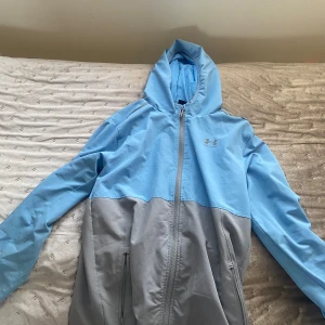 Blå och grå träningsjacka från Under Armour - Snygg träningsjacka från Under Armour i ljusblått och grått. Jackan har huva, dragkedja framtill och två sidofickor. Perfekt för dig som gillar sportig stil och vill ha något lätt att slänga på sig.