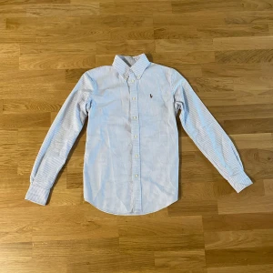 Randig skjorta från Ralph Lauren - Hej Säljer en stilren blå- och vitrandig skjorta från Ralph Lauren i storlek XS. Skjortan har långa ärmar och en klassisk krage. Den är broderad med Ralph Laurens ikoniska logga på bröstet. Perfekt för en snygg och avslappnad look. Längd= 63cm Axelbredd= 39cm armlängd= 61cm