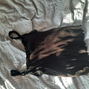 Svart ribbad linnetopp med spets - Supersöt svart ribbad linnetopp med tunna axelband och spetsdetaljer längs kanterna. Liten rosett framtill för extra cute vibe. Perfekt att styla med jeans eller kjol för en chill look.