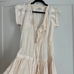 Romantisk kortklänning från Hunkydory - Supersöt ljus kortklänning från Hunkydory i tunn bomull med spetsdetaljer, knappar framtill och volangkant. Klänningen har korta ärmar med spets och broderade detaljer, perfekt för en bohemisk look. Lätt och luftig med v-ringning och knytband.