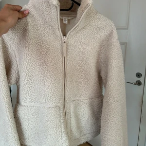 Beige teddyjacka i XS med dragkedja - Supermjuk och fluffig beige teddyjacka i storlek XS. Jackan har hög krage, hel dragkedja framtill och två stora fickor. Perfekt för kyliga dagar när du vill vara både varm och snygg. Enkel att matcha med det mesta och ger en chill vibe.