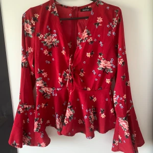 Röd blommig blus - Supersöt röd blus från Boohoo med blommigt mönster i rosa och grönt. Blusen har v-ringning med snörning framtill och långa trumpetärmar som ger en extra cool vibe. Perfekt för dig som vill sticka ut lite extra med färg och mönster.