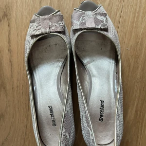 Beige spetsiga pumps med rosett från Graceland - Säljer ett par eleganta beige pumps från Graceland med spetsdetaljer och en söt rosett framtill. Skorna har öppen tå och en feminin form, perfekta för dig som vill ha en romantisk och stilren look.