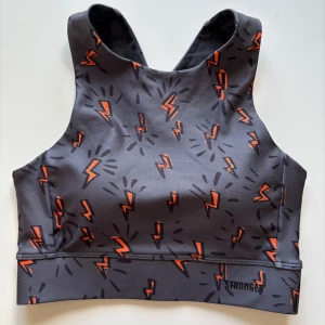 Stronger grå sporttopp med blixtmönster i nyskick storlek XS. Tights medföljer utan extra kostnad. - Stronger sporttopp i grått med coolt orange blixtmönster över hela tyget. Modellen har brottarrygg med korsade axelband och bred resår nedtill för extra stöd. Tillverkad i återvunnen polyester och elastan, perfekt för träning och aktiva dagar. Nyskick! Storlek XS. Tights i storlek XS.medföljer utan extra kostnad - har en lagning på höger knä.