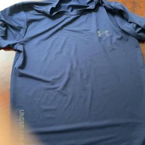 Blå träningst-shirt från Under Armour - Blå t-shirt från Under Armour med diskret logga på bröstet och tryck längs nederkanten. Perfekt för träning, tillverkad i lätt och snabbtorkande polyester. Klassisk passform och korta ärmar.