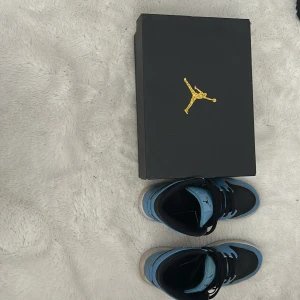 Nike Air Jordan 1 Mid Blå/Svart - Säljer ett par Nike Air Jordan 1 Mid i blått och svart med klassisk Jordan-logga på sidan. Skorna har snörning, vit sula och är tillverkade i skinn och syntet. Perfekta för dig som vill sticka ut med en ikonisk sneaker med cool färgkombo.