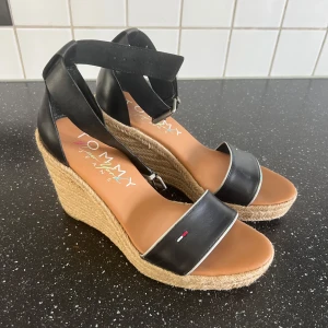 Svarta sandaler med kilklack Tommy Jeans - Snygga svarta sandaler från Tommy Jeans med hög kilklack i rep och öppen tå. Remmar i skinnimitation och justerbart spänne runt vristen. Perfekta för sommaren med stilren design och diskret logga på remmen framtill.