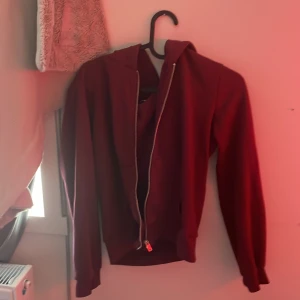 Mörkröd hoodie med dragkedja - Säljer en mörkröd hoodie med dragkedja framtill och huva. Tröjan har långa ärmar och ribbade muddar vid ärmslut och nederkant. Perfekt för dig som gillar en enkel och stilren look. Fickor framtill för extra chill vibe.