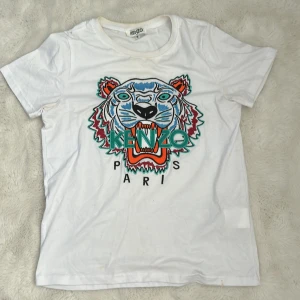 Vit Kenzo t-shirt med tigertryck - Vit t-shirt från Kenzo med ikoniskt broderat tigerhuvud och färgglada detaljer framtill. Klassisk passform, rund hals och korta ärmar. Tillverkad i 100% bomull för en skön och luftig känsla. Perfekt för dig som vill sticka ut med en statement-tee.