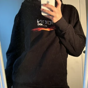 Svart hoodie  - Säljer en svart hoodie från AGON by AOC med tryck framtill och färgglada grafiska detaljer på ryggen. Tröjan har huva med dragsko och en klassisk känguruficka. Perfekt för gaming eller chill. Vunnen på dreamhack i Jönköping.