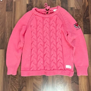 Rosa stickad tröja med broderad ros - Säljer en rosa stickad tröja med flätstickat mönster framtill och snörningsdetalj i halsen. På ena ärmen finns ett broderi med en ros och texten 'Rise Above'. Tröjan har långa ärmar och ribbade muddar.