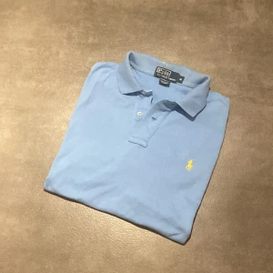 Ljusblå pikétröja från Polo Ralph Lauren - Säljer en klassisk ljusblå pikétröja från Polo Ralph Lauren med gul broderad logga på bröstet. Tröjan har korta ärmar, krage och knappar framtill. Perfekt för en stilren och avslappnad look. Den har några små fläckar på kragen (syns på sista bilden) Men ingenting illa! Bara att höra av sig vid funderingar😁👍