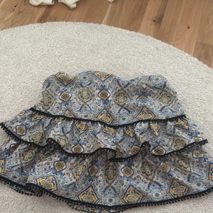 Mönstrad volangkjol med svarta detaljer - Supersöt kortkjol med blått, gult och vitt mönster samt volanger. Kjolen har svarta spetsdetaljer längs volangkanterna och är perfekt för dig som gillar färg och mönster. Bekväm passform och snygg till både sneakers och boots.