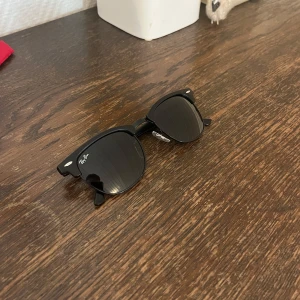 Svarta solglasögon från Ray-Ban - Snygga svarta solglasögon från Ray-Ban med klassisk clubmaster-form och mörka glas. Bågarna är i svart plast med silvriga detaljer vid gångjärnen. Perfekt accessoar för soliga dagar. Nästan sprillans nya endast använda ett fåtal gånger. Skriv vid frågor mm mvh.