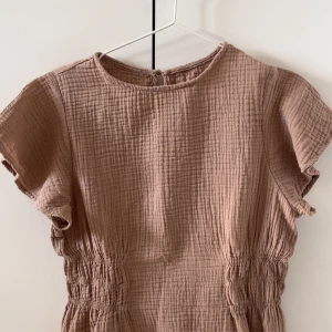 Beige blus med resår i midjan - Säljer en beige blus med korta ärmar och rund halsringning. Blusen har en avslappnad passform och resår i midjan som ger en fin siluett. Tillverkad i ett mjukt, strukturerat bomullstyg som känns skönt mot huden. Med fina volang armar. Den är mycket populär och snygg till sommaren och finare tillställningar. Använd TVÅ gånger!!💖💖🥰