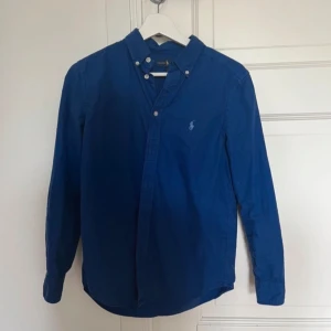 Blå skjorta från Ralph Lauren - Klassisk blå skjorta från Ralph Lauren med button-down krage och broderad logga på bröstet. Skjortan har långa ärmar och knäppning framtill. Perfekt för en stilren och tidlös look.