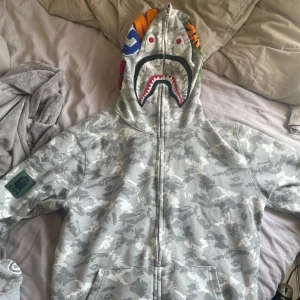 Bape hoodie (grå camo) - Säljer en grå camo hoodie från BAPE med dragkedja och ikonisk hajdesign på luvan.  Storleken är L men passar med en oversized look på mig som är xs/s, säljer pga ingen användning, varan är i fint näst intill ny skick