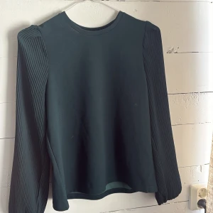 Mörk grön blus  med plisserade ärmar - Säljer en mörk grön blus med rund halsringning och långa plisserade ärmar. Toppen har en rak passform och är tillverkad i ett mjukt material som känns bekvämt mot huden. Perfekt att styla till både jeans och kjol. Endast använd fåtal gånger 