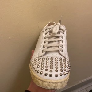 Vita sneakers med nitar från Christian Louboutin - Säljer ett par vita sneakers från Christian Louboutin med silverfärgade nitar över tån och sidorna. Skorna har klassisk röd sula, snörning och rund tå. Perfekt för dig som vill sticka ut med en edgy och lyxig stil. Dom är lite smutsiga i bilden men du får dom hellt vita och ser ut som nya