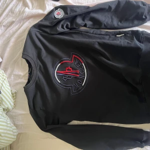 Svart sweatshirt från Moncler - Säljer en svart sweatshirt från Moncler med stort broderat logotypmotiv på bröstet och en mindre logotyp på ärmen. Tröjan har rund halsringning och långa ärmar med ribbade muddar. Perfekt för en stilren och sportig look.