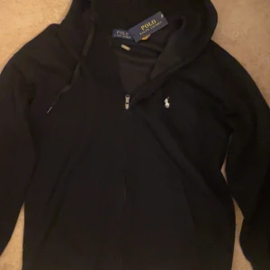 Svart hoodie med dragkedja från Polo Ralph Lauren - Svart hoodie från Polo Ralph Lauren med dragkedja framtill och klassisk vit logga på bröstet. Tröjan har huva med snörning och är tillverkad i mjukt material. Perfekt för en avslappnad och stilren look.