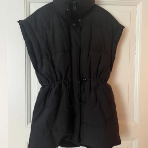 Svart dunväst GinaTricot - Svart dunväst med hög krage och dragsko i midjan. Västen har knappar och dragkedja framtill samt två stora fickor på sidorna. Perfekt för lager-på-lager och en stilren look.
