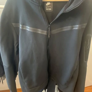 Nike Tech Kofta Svart - Säljer en svart hoodie från Nike med dragkedja och huva. Jackan har en diskret logga på bröstet och en blank rand över bröstet. Perfekt för dig som gillar sportig stil och vill ha något bekvämt till vardagen.