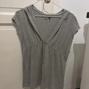 Grå kortärmad topp med huva Wearit - Grå kortärmad topp från Wearit med djup v-ringning och huva. Mjuk och skön bomullskänsla, lätt rynk under bysten och enkel knytning framtill. Perfekt för en chill och avslappnad stil.