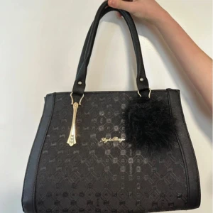 Svart handväska med mönster och pälsdetalj - Snygg svart handväska från Style Bags med ett diskret geometriskt mönster. Väskan har två handtag och en dekorativ pälsboll. Insidan är rymlig med flera fack och dragkedja för enkel organisering. Perfekt för att bära dina dagliga nödvändigheter med stil.