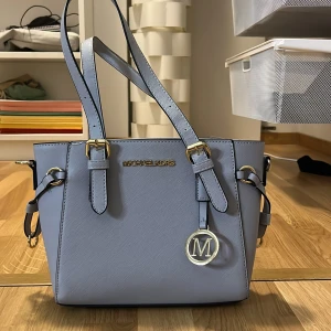 Ljusblå handväska från Michael Kors - Snygg ljusblå handväska från Michael Kors med gulddetaljer och en dekorativ M-berlock. Väskan har dubbla handtag och stilrena linjer, perfekt för en chic look. Helt ny, aldrig använd.
