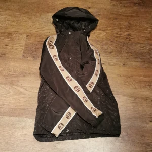 Svart jacka med huva från Gucci - Snygg svart jacka från Gucci med huva och beige detaljer längs ärmarna. Jackan har ett diskret mönster och knappar framtill. Perfekt för en stilren look.