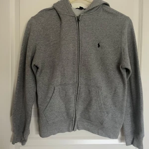 Ralph Laurent zip-up Grå - Säljer en klassisk grå hoodie från Ralph Lauren med dragkedja och huva. Tröjan har långa ärmar och två praktiska fickor framtill. I mycket bra skick. Storleken är XL i barnstorlek vilket motsvarar ca S