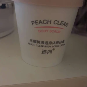 Peach Clear Body Scrub - Peach Clear Body Scrub Cream i en vit burk med rosa lock. Produkten är designad för att exfoliera och mjukgöra huden med en fräsch doft av persika. Perfekt för att ge huden en silkeslen känsla.