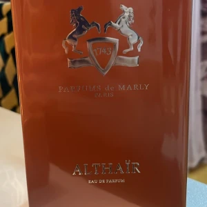 Parfums de Marly Althair Eau de Parfum - Exklusiv parfym från Parfums de Marly, Althair Eau de Parfum, med en distinkt amberdoft och varma noter av trä och vanilj. Förpackningen är elegant i en djup röd färg med märkets ikoniska emblem av två hästar och ett sköldmärke. Perfekt för den som söker en sofistikerad och tidlös doft.