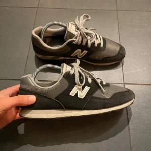 Grå och svarta sneakers från New Balance - Snygga grå och svarta sneakers från New Balance med klassisk design. Skorna har snörning och en bekväm sula, perfekt för vardagsbruk. De har en stilren look med New Balance-loggan på sidan.