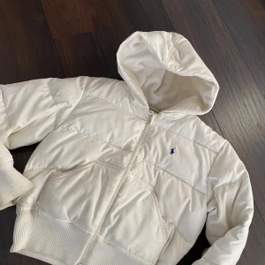 Vit dunjacka från Ralph Lauren - Säljer en snygg vit dunjacka från Ralph Lauren med huva och dragkedja. Jackan har en broderad logga på bröstet och ribbade muddar. Perfekt för kyliga dagar. 
