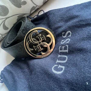 Svart bälte från Guess - Snyggt svart bälte från Guess med en stor, rund guldfärgad logotyp som spänne. Bältet har en elegant textur och kommer med en praktisk förvaringspåse. Perfekt för att ge en stilren touch till din outfit.