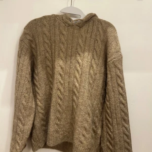 Beige stickad tröja från Gina Tricot - Mysig beige stickad tröja från Gina Tricot med kabelstickat mönster. Tröjan har en huva och långa ärmar, perfekt för kyliga dagar. Den är tillverkad i ett mjukt material som ger en skön känsla. Passar utmärkt för höst och vinter.