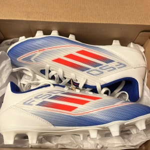 Adidas F50 Club FxG fotbollsskor - Snygga Adidas F50 Club FxG fotbollsskor i vitt med blå och röda detaljer. Skorna har en modern design med snörning och är perfekta för spel på fasta underlag. De är lätta och bekväma, vilket ger bra kontroll och hastighet på planen.