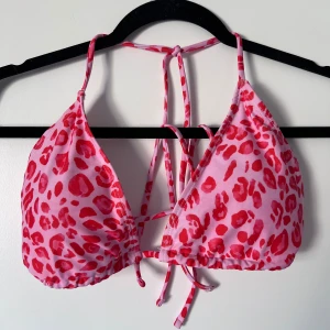 Rosaröd leopardmönstrad bikiniöverdel - Trendig rosa bikiniöverdel med rött leopardmönster. Den har knytband i nacken och ryggen för justerbar passform. Perfekt för en dag på stranden eller vid poolen. Säljer pga att den är för stor för mig. Aldrig använd!! Önskar du veta exakta mått? Tveka inte att höra av dig!