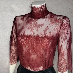 Vinröd spetsblus - Elegant vinröd spetsblus med hög krage och trekvartsärmar. Blusen har ett vackert blommigt mönster och är perfekt för en stilfull look. Den är tillverkad i ett genomskinligt spetsmaterial som ger en sofistikerad touch.