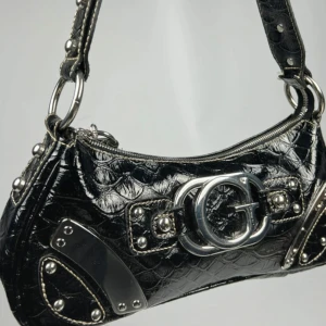 Svart Guess Y2K axelväska med metallisk logga - Snygg svart äkta vintage Guess axelväska i syntetmaterial med en stor metallisk logga framtill. Väskan har en dragkedja och dekorativa nitar som ger en cool look. Perfekt för att ge din outfit en edgy touch.