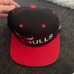 Svart och röd keps från Adidas - Snygg svart och röd keps från Adidas med Bulls-logga framtill. Kepsen har en böjd skärm och justerbar passform. Perfekt för sportiga tillfällen eller som en stilren accessoar.
