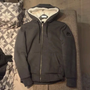 Grå hoodie från Moose Knuckles - Säljer en snygg grå hoodie från Moose Knuckles med dragkedja och huva fodrad med mjuk päls. Jackan har långa ärmar och ribbade muddar. Perfekt för kyliga dagar. 🐻
