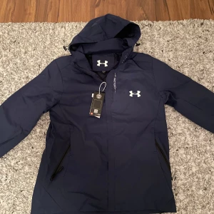 Mörkblå jacka från Under Armour - Säljer en mörkblå jacka från Under Armour med huva och dragkedja. Jackan har långa ärmar och två fickor med dragkedjor. Perfekt för utomhusaktiviteter och har en stilren design med logotyp på bröstet. Passar som M